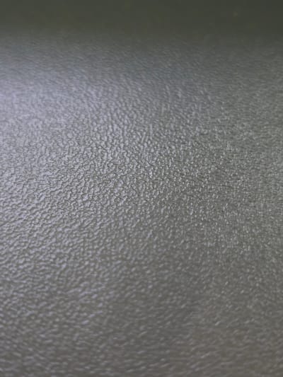 The image shows a close-up, abstract view of a textured surface. The texture is granular and bumpy, with an uneven pattern of raised and recessed areas. The color is a muted, desaturated greenish-gray, with subtle variations in shade that suggest a slight sheen or subtle lighting. The focus is shallow, with the foreground appearing sharper than the background, which gradually blurs. No specific objects, people, or actions are discernible. The image does not provide any information about its location in the city of Kotiv, Ukraine.