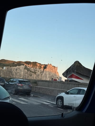 The image depicts a view through a car window, likely in Étretat, France, with the iconic white cliffs of the Alabaster Coast visible in the background. The cliffs are illuminated by the warm light of what appears to be a sunset or sunrise, casting a golden hue on their rugged surfaces. In the foreground, several cars are parked or driving on a road adjacent to a stone wall. One car, a dark-colored SUV, is emitting a plume of exhaust smoke. To the right of the image, a distinctive building with a thatched roof and red and white triangular accents is situated near the cliffs. The sky is clear and a pale blue, suggesting fair weather. The scene is captured from a low vantage point, emphasizing the perspective from within a vehicle.