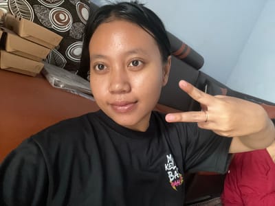 This is a selfie of a person indoors, likely at home, given the casual setting with furniture and a patterned cushion. The person is wearing a black t-shirt with text on it. The text on the t-shirt reads "MI KEL BA RAH" with some stylized graphic elements above it. The person is making a peace sign with their left hand, which has a thin ring on one finger. The overall mood appears relaxed and informal. There are no clear indicators of the time of day or weather. The presence of small cardboard boxes stacked on a patterned cushion suggests some form of organization or preparation, but no specific activity is definitively shown. The background includes what appears to be a piece of red fabric and furniture.