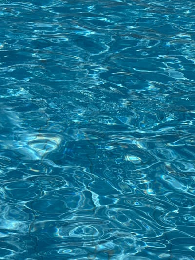 The image shows the surface of a swimming pool in Bat Yam, Israel. The water is a clear blue, and the sunlight creates patterns on the bottom of the pool. The tiles of the pool are visible through the water. No people or other objects are visible.