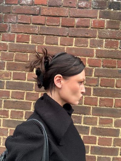 A woman is standing in front of a brick wall. She is wearing a black sweater and her hair is in a messy bun. The wall is made of red bricks with light-colored mortar. It is not possible to determine the location of the image in the city of Wealden, United Kingdom, or any additional details from the image provided.