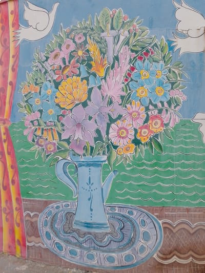 This is a ceramic mural located on the exterior wall of the Public Market of Cuiabá, Brazil. The artwork depicts a vibrant, stylized floral arrangement in a blue pitcher, placed on a decorative blue and grey platter. The flowers are rendered in a variety of bright colors, including pink, blue, yellow, orange, and purple, with green leaves and accents. Behind the bouquet, a light blue background suggests a sky, with two white doves in flight, one on the upper left and another on the upper right. A red and yellow curtain is visible on the far left of the mural. The foreground shows a textured brown surface with a wavy pattern, and green wavy lines representing water.