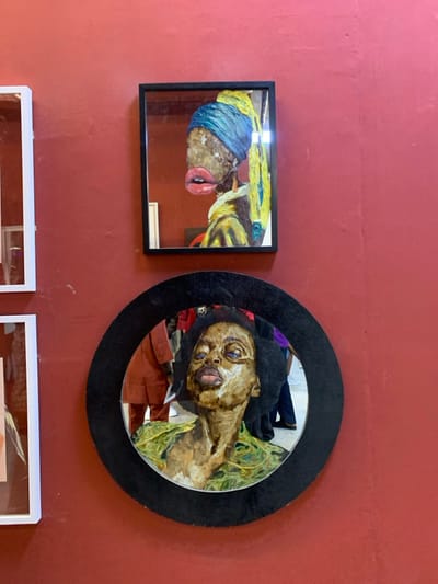 The scene displays a reddish-brown textured wall in Itirin, Nigeria, serving as an exhibition surface for framed artworks. Centrally positioned in the upper half is a rectangular black-framed artwork, depicting a stylized, mixed-media rendition of "Girl with a Pearl Earring." This artwork features a figure with dark, textured skin, exaggerated red lips, an elongated ear with an earring, a blue and yellow head covering, and a yellow garment. Below it, slightly to the right, is a larger circular artwork with a thick, textured black frame. This piece presents a stylized bust of an African woman with dark, curly hair, a light brown facial texture with prominent features, and eyes gazing upward; she appears to wear a green patterned garment. Both artworks utilize reflective surfaces. The circular frame clearly reflects two individuals in the background: one partially visible on the left wearing a reddish-brown garment, and another on the right in dark pants and a visible purple top. Two white-framed objects are partially visible along the left edge of the image.