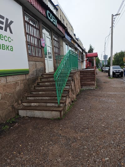 The image shows a street scene in Kumertau, Russia, featuring the entrance to a commercial building with a prominent "CDEK" logo. The building has a stone facade and large windows. A green metal staircase with a handrail leads up to the entrance. To the left of the building, a large white sign with green lettering is visible, part of which reads "пресс- доставка" (press-delivery). A red awning covers a section of the building to the right. The street in front of the building is paved with asphalt and gravel. Several cars are parked along the street, and a street light pole is visible. The sky is overcast, indicating a cloudy day.