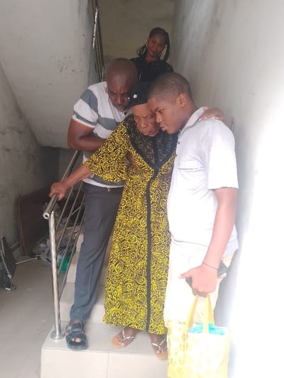 In a building located in Mgbuoshimini, Nigeria, four individuals are positioned on a concrete staircase. An elderly woman, centrally located on a step, wears a long yellow and black patterned dress, a black headscarf, and light-colored open-toe sandals. She is being assisted down the stairs by two men. To her left, an adult male with a bald head, wearing a striped polo shirt and dark trousers, supports her with his left arm around her back. His right hand rests on a silver metal handrail. To her right, a young male, wearing a light grey collared shirt and light trousers, supports her with his left arm around her shoulder. He holds a yellow bag with blue lining in his right hand. The elderly woman's right hand is also on the metal handrail. In the upper background, an adult woman with dark braided hair, wearing a dark top, observes from a higher position on the staircase. The concrete walls and steps are light-colored. Various items, including a brown board, are visible as clutter below the handrail on the left side of the stairs. Natural light illuminates the scene, primarily from the right.