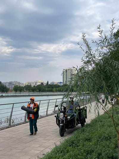 The image depicts a cloudy day scene along a waterfront promenade in Almaty, Kazakhstan. In the foreground, a male city worker in an orange cap and green work uniform walks on a paved path, carrying a black cylindrical object. To his right, another man is driving a three-wheeled electric vehicle. A metal railing separates the promenade from a body of water. In the background, several multi-story buildings are visible, along with trees and a distant shoreline. A large weeping willow tree with its branches hanging down frames the right side of the image. The overall setting suggests a public park or recreational area within the city.