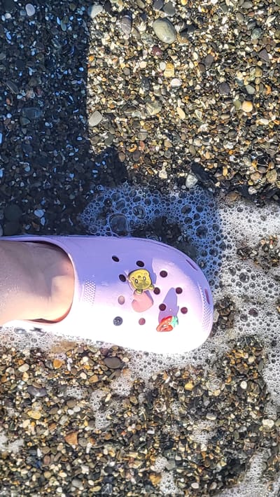 A person's foot, partially submerged in shallow water, is wearing a light pink Croc shoe. The shoe is adorned with two charms: a yellow bear and a red strawberry. The foot and shoe are positioned on a beach covered in numerous small, multi-colored pebbles. Gentle waves are washing over the pebbles and the shoe, creating a line of white foam and bubbles. The scene is outdoors, likely during the daytime, given the bright lighting and the shadow cast by the shoe. The overall impression is one of a relaxed, casual moment at a beach.
