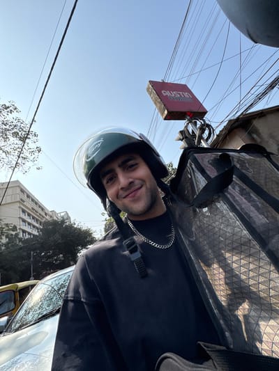 A man wearing a helmet smiles at the camera, with a delivery bag strapped to his back. The scene is captured from a low angle, showing power lines and a sign reading "Austin Plywood" above. In the background, trees and a tall building are visible. The lighting suggests it's daytime. The man is wearing a dark-colored shirt and a silver chain is visible on the side of his face. Part of a car or autorickshaw can be seen on the right side of the frame.