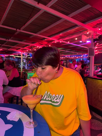 A woman in a yellow "Money" shirt sips a yellow drink with a straw at a table at a bar. Another woman sits nearby in a black tank top and white skirt. Behind them, other patrons are visible in the crowded, indoor/outdoor bar. The bar has a bamboo-lined roof structure crisscrossed with red and pink string lights. This scene is located in Canggu, Indonesia.