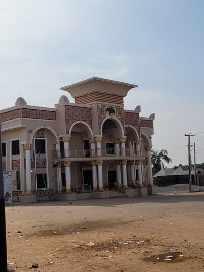 The image depicts a two-story traditional palace building, identified by signage as "AREN EGON'S PALACE," located in Nassarawa Egon, Nigeria. The structure is characterized by a cream-colored facade with extensive brown and gold geometric patterns and trim. It features a central entrance with a set of steps leading to a raised ground floor and a first-floor balcony. The entrance and balcony are supported by multiple white columns with gold capitals and bases, framed by ornate arches. Above the central arch on the first floor, a dark silhouette of an elephant is depicted. Two flags, one appearing to be the Nigerian national flag, are mounted on poles near the left side of the main entrance. Dark-paned windows are visible on both levels.

The foreground consists of an unpaved, reddish-brown dirt area, heavily littered with various pieces of paper, plastic, and small debris. No individuals or vehicles are present in the immediate foreground. To the right of the palace, in the background, utility poles with power lines are visible, along with a prominent palm tree. Further back, residential structures with corrugated metal roofs are discernible behind a low wall, indicating a populated area. The sky above is clear and light blue, suggesting daylight conditions.