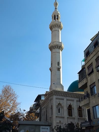 An urban exterior scene in Sarouja Municipality, Syria, depicts a prominent mosque minaret and dome under a clear blue sky. The central subject is a tall, slender minaret constructed of light-colored stone, featuring an octagonal shaft, intricate decorative carvings, and two distinct balconies with balustrades. The minaret culminates in a pointed, onion-domed finial. Partially visible behind the minaret and to its right is a large, dark green or teal dome, indicating the main structure of the mosque. Adjacent to the mosque on the right is a multi-story building, appearing to be residential or commercial, constructed from light brown or beige materials. This building exhibits multiple balconies, windows, some with dark awnings, and air conditioning units. Arabic script is discernable on a sign or awning at the street level of this building. In the lower-left foreground, a grey utility box with "SHEIN" written in white is partially visible, positioned near a deciduous tree with sparse, golden-brown autumnal foliage. A single black utility wire stretches horizontally across the mid-ground. The scene is illuminated by bright daylight, and no individuals are present.