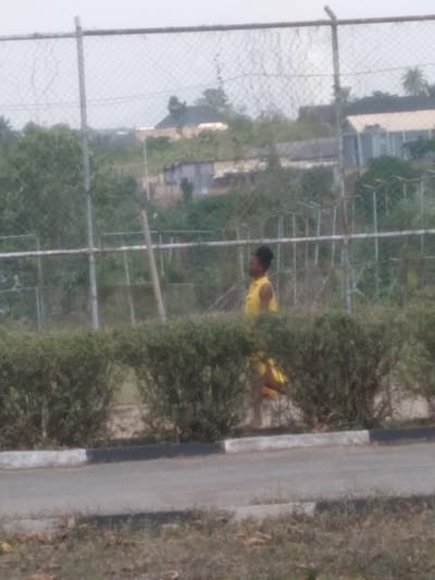 This image depicts an outdoor scene in Osu, Nigeria. A single individual with dark skin and short, dark hair, wearing a bright yellow top and matching yellow shorts, is captured in a running or brisk walking posture, moving from left to right. The person's lower legs are partially obscured by a horizontal line of dense, green, trimmed hedges. In the foreground, a paved surface with a white and black painted curb is visible, bordered by dry grass and sparse vegetation. Behind the hedges, a metal chain-link fence with vertical support posts extends across the mid-ground. The background reveals an expanse of green foliage, and several distant buildings are discernible against a light, overcast sky; these include a multi-story building with a dark roof on the left and a lighter-colored building on the right.
