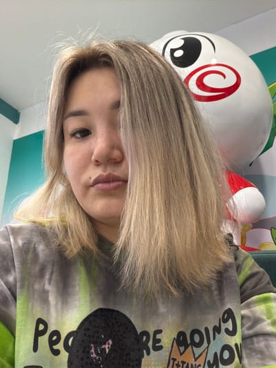 The media file is a selfie of a young woman with blonde hair, taken indoors. She is wearing a tie-dye shirt with green and grey tones. The shirt has text printed on it, which appears to say "Peace" and "Going". The background is a wall with turquoise and white patterns, and a large white inflatable object with a red swirl design and black eyes is visible behind her head. The lighting suggests it is daytime. The woman's expression is neutral. The location context is Astana, Kazakhstan.
