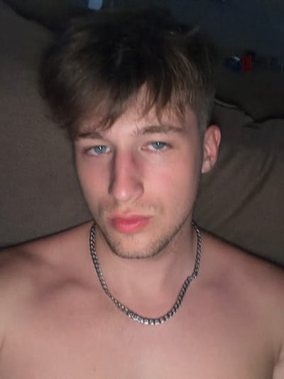 The image is a close-up, waist-up, selfie-style photograph of a young Caucasian male with light skin, light brown hair, and blue eyes. He is shirtless and wearing a silver chain necklace. His lips are slightly parted, and his gaze is directed at the camera. The background is dark and appears to be indoors, possibly a couch or upholstered furniture, with some out-of-focus objects in the distance. There is no discernible indication of the location being Vanino, Russia.