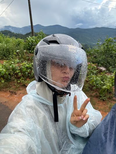 A person is shown in a close-up shot, wearing a helmet with a clear visor covered in raindrops. The person is also wearing a light blue raincoat, which appears to be made of plastic. Their right hand is raised, forming a peace sign with manicured fingernails. The background shows a mountainous landscape with lush green vegetation under a cloudy sky, suggesting a rural or natural setting. The ground appears to be wet and muddy. The context provided states the location is Nao Sẻ, Vietnam, which is a rural district in the Tuyen Quang province of Vietnam, known for its mountainous terrain.