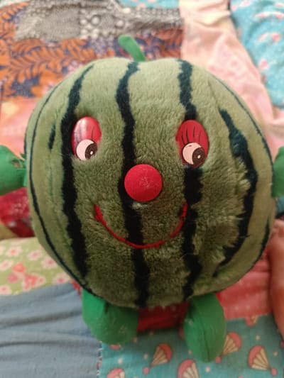 A close-up, top-down shot shows a plush watermelon toy with a smiling face, set against a background of colorful, patterned fabrics. The toy has a round green body with black stripes, two red eyes with black pupils and eyelashes, a red round nose, and a red stitched smile. It has small green arms and legs, and a small green stem on top. The fabric background features a patchwork of different patterns, including floral designs, abstract prints, and a light blue fabric with parachute illustrations. There are no visible people or specific landmarks to indicate a location in the city of Nesvizh, Belarus. The lighting is moderate, suggesting an indoor environment.