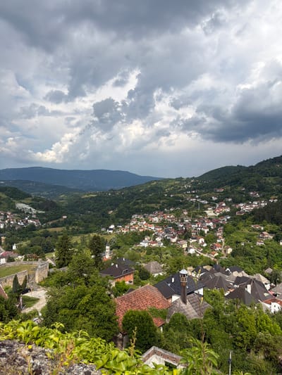 This media file shows a panoramic view of a hillside town, likely Jajce, Bosnia and Herzegovina, nestled amidst lush green trees and rolling hills. The town is characterized by its densely packed houses with red-tiled roofs, interspersed with smaller buildings and narrow pathways winding through the landscape. The architecture suggests a historical European setting.
The scene is set under a dramatic sky filled with heavy, dark clouds, indicating overcast or impending stormy weather. The lighting is diffused, casting a soft glow on the scene. The time of day appears to be afternoon or late morning, given the cloud cover.
In the foreground, a stone wall or fortification is visible, overgrown with vibrant green ivy and other vegetation, suggesting an ancient structure. A few sparse branches with leaves are also present in the immediate foreground. Beyond the wall, the view opens up to reveal the town cascading down the slope, with a prominent church spire visible among the buildings. Further in the distance, a large, dark mountain range dominates the horizon, shrouded in a slight haze.
There are no discernible people or specific activities taking place. The focus is on the natural landscape and the architectural elements of the town, creating a sense of timelessness and tranquility, despite the brooding sky. The overall mood is serene and slightly dramatic due to the weather. No visible text is present in the image.