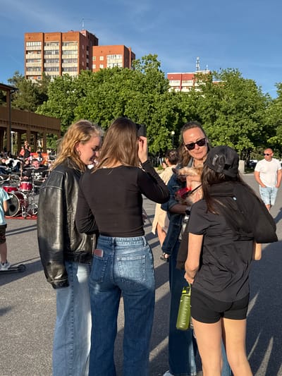A group of young people are gathered outdoors in a paved area, possibly a public square or park, in Tallinn, Estonia. The background features multi-story brick buildings and lush green trees. In the foreground, four individuals are the primary focus.

On the left, a woman with blonde, wavy hair wears a black leather jacket over a light blue denim shirt and light blue wide-leg jeans. She is looking down and to her right. Next to her, a woman with brown hair, wearing a black long-sleeved shirt and blue jeans, is facing away from the camera, also looking down. A pink object, possibly a wallet or phone case, is visible in her back jeans pocket.

To the right of these two women, a third woman with dark hair, sunglasses, and a denim jacket is holding a small, tan-colored dog. She is looking at the dog. A fourth person, wearing a black baseball cap and a black hoodie over a black t-shirt, is facing the woman with the dog, holding a green water bottle. Their back is mostly to the camera.

In the background, a drum set is visible, suggesting a possible outdoor concert or rehearsal. Other people and a man in a white t-shirt and shorts are also present in the middle ground and background. The ground is asphalt, and sunlight casts shadows, indicating it is daytime.