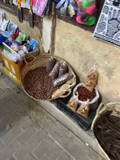 This image depicts a market stall, likely within a souk in the Medina of Fez, Morocco. The scene presents a static display of various goods, with no individuals present.
The foreground features a worn, light-colored tiled or paved floor. Against a weathered, light brown wall, several items are arranged for sale. On the ground level, starting from the left, a yellow cardboard box holds an assortment of small, colorful plastic and fabric items. Adjacent to it, a large, round woven basket is filled with reddish-brown legumes or nuts, with several smaller, clear plastic bags containing the same product nestled within. To the right, a white sack or container on a black plastic crate holds a quantity of small, dark reddish-brown dried fruits or berries. Four clear plastic bags, standing upright, contain a fine reddish-brown powder, consistent with ground spices or henna, with indistinct Arabic script visible on their labels. Further to the right, a partial view of another woven basket reveals dark brown, fibrous material, possibly dried date strands or other dried food products.
The wall directly above the displayed goods exhibits various items hanging from it. These include colorful plastic pouches or accessories on the left, two black-handled foot files in the center-left, and a clear plastic bag containing green and pink spherical objects in the upper-right. A black-and-white poster or sign featuring intricate patterns and Arabic calligraphy is also affixed to the wall on the far right. The wall itself shows signs of age and wear, consistent with an active, historical marketplace environment.