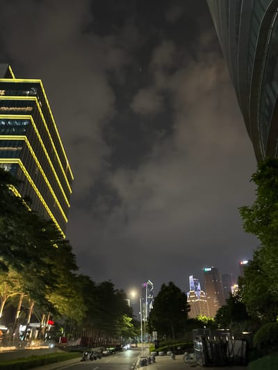 A nighttime street scene in Haizhu District, China, features a modern cityscape. On the left, a multi-story building is illuminated with yellow lights outlining its floors. To the right of this building, trees with glowing lights are planted along a sidewalk. Further down the street, a road is lined with parked motorcycles and a few cars. In the distance, several tall skyscrapers are lit up, some with distinctive colored lights. The sky is dark and cloudy. A portion of a curved building is visible on the far right.