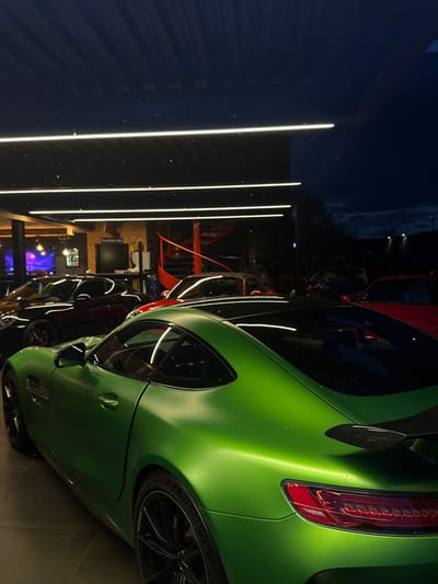 The image displays the interior of a car showroom or garage in Souffelweyersheim, France, during nighttime. In the foreground, a matte green Mercedes-AMG GT R sports car is prominently featured, positioned facing slightly left and away from the viewer. Its surface is speckled with small water droplets. The vehicle has black multi-spoke wheels and a carbon fiber rear spoiler.
Within the illuminated interior, behind the green car, multiple other high-performance sports cars, predominantly black, dark grey, and red, are parked. The ceiling is equipped with several bright, horizontal LED strip lights. To the right in the background, a distinctive red spiral staircase ascends. A large monitor displaying "Porsche.de" is visible on a wall. Beyond the glass facade of the building, the exterior scene is dark, characteristic of night, with faint distant lights and silhouetted foliage against a dark blue-black sky. No individuals are present in the visible area.