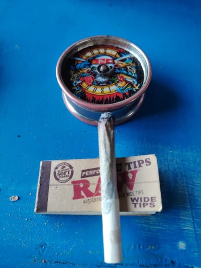 The image captures a close-up, eye-level view of several items arranged on a vibrant blue, textured surface in Ksar es-Seghir, Morocco.
In the upper central part of the frame, a metallic, circular object, likely a grinder or an ashtray, displays a colorful Guns N' Roses band logo on its top surface. The logo features a skull wearing a bandana emblazoned with "N' ROSES," flanked by feathered wings, roses, and banners spelling out "GUNS" and "ROSES." The object has a copper-colored rim and a silvery body.
Standing upright in front of this object is a hand-rolled cigarette (joint). Its tip is visibly burnt and topped with a small amount of white ash, indicating it has recently been lit or is currently smoldering. The joint itself is wrapped in light-colored paper.
Positioned below the joint and extending towards the bottom right is a small, rectangular cardboard box of "RAW" brand rolling tips. The box, in a natural light brown color, clearly displays the text "RAW" in red, along with "PERFORATED WIDE TIPS," and smaller inscriptions such as "AUTHENTIC GENUINE SOFT FIBERS ORIGINAL."
The blue surface beneath the items appears slightly worn and has a few small dark specks, possibly ash or debris. The scene suggests a casual moment of preparation or relaxation.