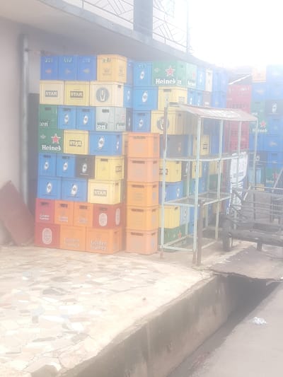 The image depicts a large stack of beverage crates, primarily beer crates, outside a building in Enugu, Nigeria. The crates are made of plastic and are in various colors including blue, yellow, green, and red. Many of the crates are labeled with brand names such as "STAR," "Heineken," and "Golden Guinea." Some of the crates appear to be empty, while others may contain bottles. The crates are stacked high, forming a wall-like structure. In the foreground, there is a mosaic tiled surface, possibly a sidewalk or patio, and a drainage ditch. Behind the crates, a metal shelving unit is visible, along with what appears to be a cart or trolley. The overall scene suggests a storage area or a point of sale for beverages. The lighting is bright, indicating it is daytime.