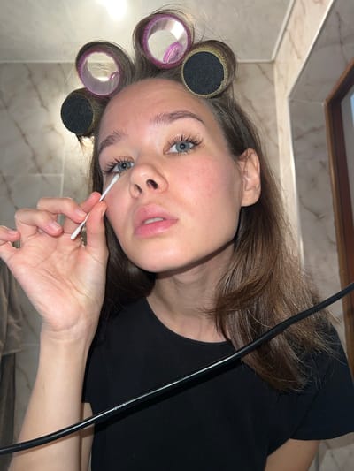The image shows a young woman with blue eyes and brown hair styled with hair rollers. She is wearing a black t-shirt and is holding a cotton swab to her left eye. Her left hand is visible, holding the cotton swab. The background appears to be a bathroom with marble-tiled walls. A black cable runs diagonally across the lower portion of the image. The lighting suggests an indoor setting. The image is a close-up shot focusing on the woman's face and upper body. There are no discernible elements in the image that can be used to ascertain the location in the city of Batumi, Georgia.