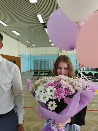 A young woman with fair skin and wavy reddish-brown hair holds a large bouquet of flowers, predominantly white and shades of pink and purple, wrapped in lavender-colored paper. Balloons in pastel shades of pink, purple, and white float above the bouquet. She appears to be smiling down at the flowers.
To her left is a man, only partially visible, wearing a white long-sleeved shirt. The setting appears to be indoors, likely a gymnasium or large hall, given the ceiling with recessed lighting and the basketball hoops visible in the background. The walls are painted in light blue and white. Chairs are lined up against the wall in the background.
There is no evidence to indicate this photograph was taken specifically in the city of Gremyachinsk, Russia.