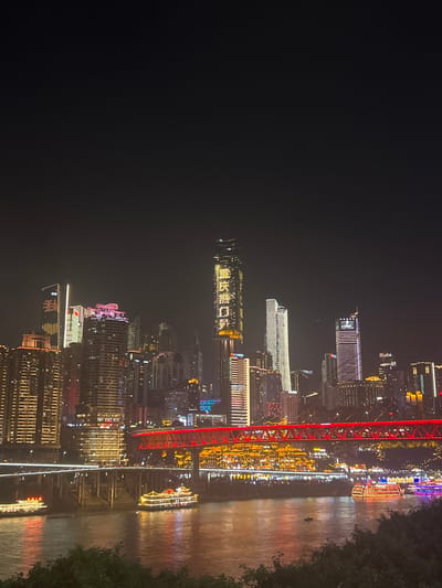 The image shows a nighttime cityscape of Jiangbeicheng Subdistrict, China, dominated by a river and illuminated skyscrapers. The river in the foreground is reflecting the lights of the city, with several boats of various sizes traversing its surface. Two larger boats, adorned with numerous lights and traditional Chinese architectural elements, are prominent. A red illuminated bridge spans across the river in the middle ground. Behind the bridge, the cityscape is a dense collection of tall buildings, many of which are lit up with colorful lights, including advertisements and patterns displayed on their facades. The sky above is dark, suggesting it is late at night. The overall scene depicts a vibrant and bustling urban environment.