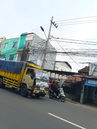 A yellow truck with a blue tarp drives down a road next to a motorcycle carrying two people. Power lines crisscross the sky above buildings of varied styles and colors. The buildings feature a mix of modern and traditional designs, including one with a teal and pink facade. A utility pole with a streetlight stands close to the truck, adding to the complexity of the overhead wires.