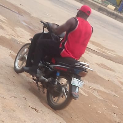An eye-level shot captures a person riding a motorcycle on a light brown, possibly dusty or unpaved road, with some lighter gray patches indicating mixed road surfaces. The image is tilted, suggesting a dynamic angle or movement.
The primary subject is a male individual, seen from behind and slightly to the left, wearing a bright red baseball cap and a red sleeveless top with white trim on the shoulders. His skin appears dark. He is seated on a black and grey motorcycle, which has a visible rear wheel, a silver-colored exhaust pipe, and a partially obscured license plate. The license plate appears to have some indistinct white characters on a dark background, with the number "1" faintly discernible. The front wheel of the motorcycle is also visible.
The background on the left is indistinct, showing faint shapes that could be distant buildings or foliage. The overall lighting suggests it is daytime. No specific time of day or weather conditions are explicitly clear, though the dusty road surface hints at dry conditions. The image does not convey any specific emotions. The location context provided is Sardauna, Nigeria.