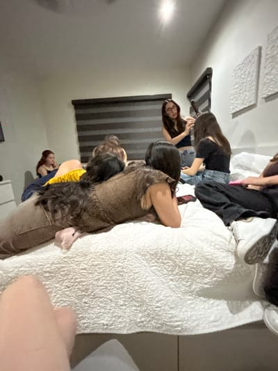 Several people are congregated on and around a bed, covered with a white, quilted bedspread. One woman is wearing a brown jumpsuit and kneeling on the bed, while another person in a black top and jeans is leaning over the bed. Two other women can be seen further back in the scene, one with long hair holding an object, and another wearing a blue shirt. The room has dark striped blinds on the window. There are two square textured panels on the ceiling. The location appears to be an interior space. The location is not discernible as Tampico, Mexico.