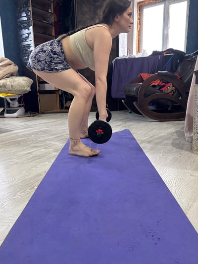An adult woman is depicted exercising indoors, likely performing a squat or deadlift variation, holding a black dumbbell with red collars. She is positioned barefoot on a purple yoga mat, which rests on a light wood-patterned floor.
The woman wears a light beige sports bra and patterned dark blue and white shorts, with her dark hair pulled back in a braid. Her expression is focused, and she gazes slightly upwards. A small, dark, possibly cursive tattoo is visible on her right ankle.
The setting appears to be a home in Rusalevka, Russia. Bright natural light streams through a large window in the background, suggesting it is daytime. Around the window, various household items and furniture are visible, including what looks like a dark wooden chair or storage unit and piled textiles. Additional indistinct objects, such as a cushion and a white appliance, are present in the lower left of the frame. The overall scene conveys a casual, home workout environment. No clearly legible text is visible other than the small tattoo.