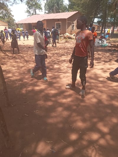 The image depicts a scene in Malava, Kenya (latitude: 0.53, longitude: 34.812).  Two young men are standing in a dusty, dirt yard, surrounded by several other individuals. The men are wearing casual clothing, and one of them is wearing a red t-shirt. They are both standing in a relaxed pose, with their feet slightly apart and their hands resting on their sides. The yard is covered in dirt and is scattered with debris. There are shadows cast by trees, and a nearby building, with a roof that is partially visible, appears to be made of concrete. The other individuals are moving and interacting, but their actions are not clear. The scene is uncluttered, with minimal background detail. The atmosphere is casual and relaxed.  The image has a general focus on the two young men in the foreground.  The image appears to have been taken during the daytime.