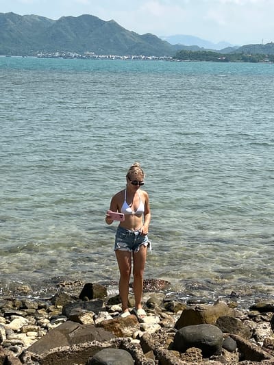 A young woman stands on a rocky shore at the edge of the sea. She is wearing a white bikini top and denim shorts. She is holding a pink smartphone in her right hand, and her left hand is in her pocket. She is looking down at the phone. Her hair is blonde and pulled up in a bun. She is wearing sunglasses. The sea is calm and clear, with visible rocks beneath the surface. In the background, there are green mountains and a distant coastline with buildings. The sky is partly cloudy. The image appears to have been taken in Ninh Ích, Vietnam, given the landscape.