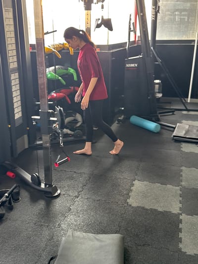 A person stands barefoot in a gym. They wear a maroon long-sleeved shirt and black leggings. The gym has black and grey floor tiles. Equipment includes a XEBEX rower, various weight machines, and a blue foam roller. The person is looking down.