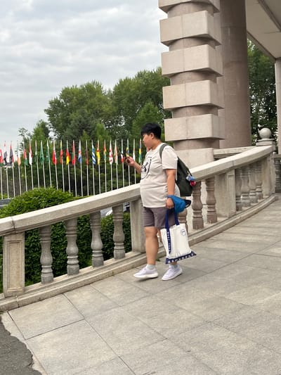 A person is standing on a stone walkway with a railing and a row of international flags in the background. The person is wearing a light-colored t-shirt, shorts, and white sneakers, and is carrying a backpack and a tote bag. They are looking down at their phone. The flags appear to be in front of a building with large pillars. This location is the Changchun World Sculpture Park, a prominent park in Changchun City, Jilin Province, China, known for its collection of sculptures and international flags representing various countries.