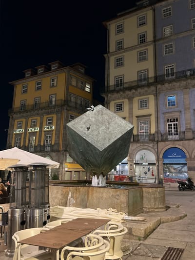 The image captures a night scene in the city of Porto, Portugal, specifically at the Praça da Liberdade. Dominating the foreground is a large, metallic cube sculpture, seemingly a fountain, with water spraying from its base. Surrounding the fountain are outdoor dining tables and chairs, some covered with white cloths. Two tall, silver patio heaters stand to the left of the frame. Behind the fountain, the Pestana Vintage Porto Hotel is visible, its yellow facade adorned with illuminated signage that reads "PESTANA VINTAGE PORTO". To the right of the hotel, another building features a light blue tiled facade. In the background, a motorcycle is parked near a poster. The scene is illuminated by streetlights and the interior lights of the buildings.