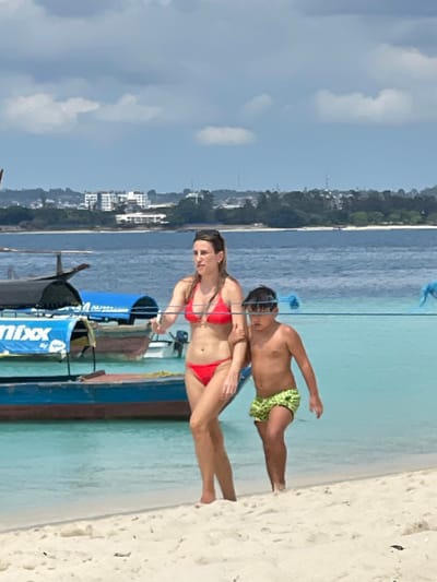 An outdoor, eye-level photograph depicts a woman and a child walking on a sandy beach in Zanzibar, Tanzania, under a partly cloudy sky. The woman, appearing to be an adult, wears a red two-piece bikini and has dark hair. She walks barefoot on the light-colored sand, looking straight ahead. To her right and slightly behind her, a young male child, shirtless and wearing green patterned swim trunks and dark-rimmed goggles on his head, walks barefoot. A blue rope or line extends horizontally across the frame at their mid-torso height. In the clear, shallow, turquoise water behind them, several boats are visible. One boat features a blue canopy with the partial text "Nixx" in white. In the distant background, across deeper blue water, a coastline with buildings and green vegetation is visible under the bright daytime sun.