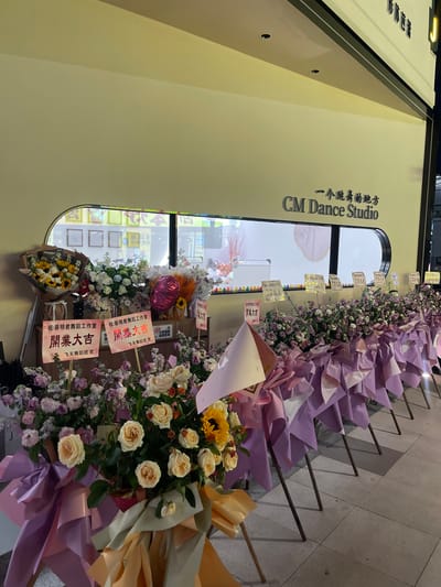 This is a medium shot, slightly tilted photograph of a storefront decorated with flowers and signage. The storefront appears to be a dance studio, indicated by the text "CM Dance Studio" and "一个跳舞的地方" (A place to dance). The studio's entrance is a large, arched window.
The foreground is dominated by several large floral arrangements, likely congratulatory bouquets for an opening. These bouquets feature a mix of roses, sunflowers, and other flowers in shades of cream, yellow, and purple, with prominent lavender and pink wrapping paper and ribbons. Some of the bouquets have small signs attached.
The sign to the left of the arched window displays Chinese characters and the name "CM Dance Studio." There are also several pink and white signs interspersed among the flowers in front of the window. Some of these signs contain Chinese text, including "祝:蔡明君舞蹈工作室" (Wishing Cai Mingjun Dance Studio) and "開業大吉" (Grand Opening Blessing/Good Fortune). One sign also mentions "飞天舞蹈团 贺" (Feitian Dance Troupe Congratulates). Another sign in the background indicates a "24小时视频 监控区域" (24-hour video surveillance area).
The overall scene suggests a celebratory event, most likely the grand opening of the dance studio. The presence of multiple flower arrangements and congratulatory signs points to a business opening or anniversary. The lighting suggests it might be evening or a dimly lit indoor space. The location context of Panyu District, China, is provided.