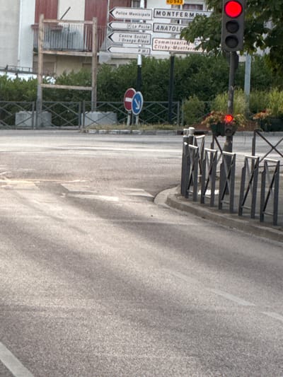 The image shows a street scene in Clichy-sous-Bois, France, with a traffic light displaying a red signal. Multiple street signs are visible, indicating directions to "Police Municipale," "MONTFERMEIL," "La Poste," "ANATOLE," "Centre Social | Orange Bleue," and "Commissariat | Clichy-Montfermeil." A metal fence separates the road from a grassy area with hedges. In the foreground, the asphalt road shows tire marks and a white line. The environment appears to be an urban intersection on a sunny day.