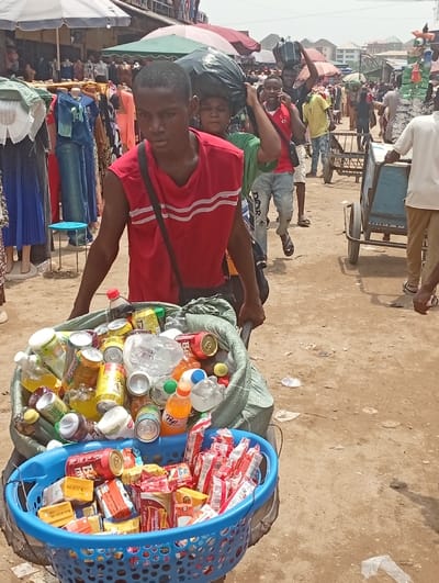 The image depicts a crowded outdoor market scene in Fegge, Nigeria, during daylight hours. In the foreground, a young male wearing a red sleeveless shirt with white horizontal stripes and a dark shoulder bag pushes a two-wheeled cart. The cart carries two distinct loads: on top, within a green woven sack, is an assortment of discarded aluminum beverage cans and plastic bottles, some containing residual liquids. Beneath this, in a blue plastic laundry basket, are various packaged snack foods, primarily biscuits and instant noodles.
Behind the foreground subject, multiple individuals navigate the market. One person directly behind him also carries a large black bag on their head. Further into the background, other people are visible, including a male in a red shirt talking on a mobile phone and another person pushing a different two-wheeled cart. Market stalls line the area, displaying clothing on mannequins, with various colored umbrellas and awnings providing shade. The ground is unpaved, consisting of dirt, with some scattered debris. Buildings are visible in the far background under a bright sky.