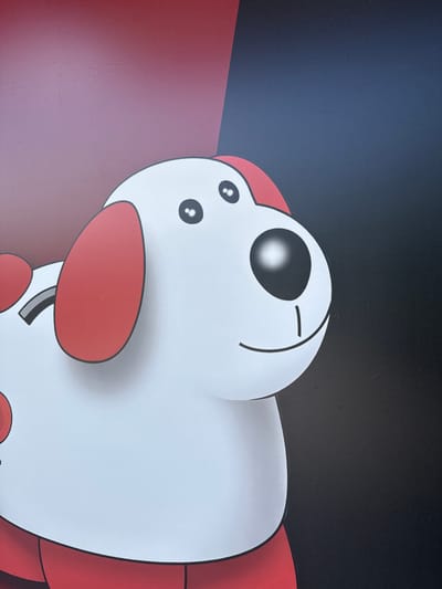 This media file is a close-up photograph of a cartoon character, likely a digital illustration, displayed on a screen. The character is predominantly white with red accents. It has large, round black eyes, with one eye appearing to be closed or squinting. Two smaller dots above the visible eye suggest nostrils or additional facial features. The character has prominent red rounded shapes on its face, possibly representing cheeks or ears, and a larger red shape below, resembling a mouth or chin. A thin black line curves from the top of the head, framing the closed eye. The character's body appears bulbous and is outlined in black. Red elements are also visible on the sides of the character's body.

The background is divided into two sections: a dark, almost black upper portion with a subtle textured pattern, and a lower section that transitions from a deep red to a lighter, pinkish-red hue. The lighting suggests the image is illuminated from the left, casting a shadow to the right of the character. There are some minor imperfections on the screen, appearing as faint scratches or dust specks. No text is visible in the image. The scene is static, showing only the character's head and a portion of its body. The overall impression is of a cute or playful character. The context of Shumen, Bulgaria, does not provide any direct visual cues within the image itself.