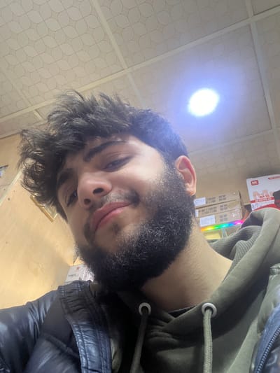 The image displays a close-up, low-angle selfie of a male individual with dark, curly hair and a full beard, looking directly at the camera with a subtle smile. He is wearing a dark, possibly navy or black, puffer jacket over an olive-green hooded sweatshirt with visible drawstrings.

The background suggests an indoor commercial or storage environment. A dropped ceiling with white, patterned panels and a single bright, circular overhead light fixture is visible. To the right of the subject, stacks of cardboard boxes are present, with partial text visible, including "Mr.", "EXTRA BASS", and "SPEAKER BAR" on one box, and "BAGA" on another. Below these boxes, a strip of multi-colored LED lights (green, blue, purple) is visible. To the left, a light-colored wooden or paneling wall is seen, featuring a small, framed object and a box partially labeled "TONER". The precise location within Sarouja Municipality, Syria, cannot be determined from the visual information provided.