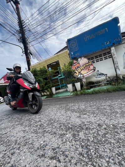A person wearing a silver helmet and a dark jacket rides a red and black scooter on an asphalt road. The scooter has a black windscreen and silver accents. The person is looking forward, with their hands on the handlebars. The road is wet, suggesting recent rain. In the background, there is a blue sign with Thai writing, a wooden pole with numerous electrical wires, a yellow building with a white trim, and some potted plants. A mural of a bird is painted on the wall of the building. The sky is overcast with white clouds. The image appears to be taken in Choeng Thale, Thailand, as indicated by the Thai writing on the sign and the overall environment.