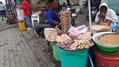 A street vendor with blue shirt stands beside stacks of packaged treats on buckets. One stack contains peanut snacks in plastic. The other has cylindrical-shaped snacks, some pinkish, others white. Beside the snack stall, a person in a white hoodie sits eating, with baskets of peanuts nearby. Other people walk around and vehicles are parked, adding to the hustle of the street scene.