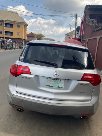 A silver Acura MDX is parked on the street in Umuode, Nigeria. The rear of the vehicle is prominently displayed, revealing a Lagos license plate. The surrounding environment includes buildings, power lines, and other vehicles. People are visible in the background.