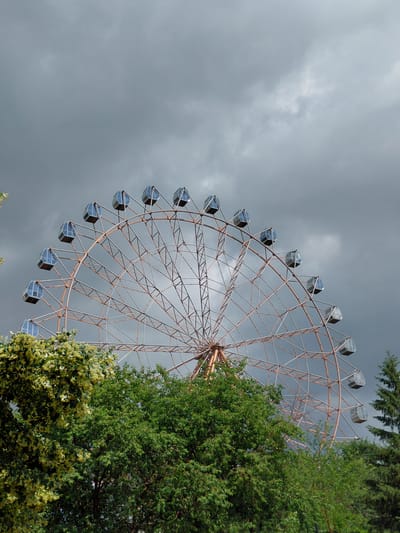 An amusement park Ferris wheel is visible against a dark, cloudy sky. The wheel has a steel frame with numerous spokes and is equipped with multiple passenger cabins, which are partially obscured by trees in the foreground. The Ferris wheel appears to be located in a park or recreational area, with dense green foliage surrounding its base. The sky is heavily overcast, suggesting impending rain or a stormy weather condition. The image does not contain any people. The Ferris wheel is approximately centered in the frame, and its upper portion extends towards the top of the image. The trees in the foreground are lush and green, with one tree on the left having lighter green or yellow foliage, possibly indicating flowering. The overall scene conveys a slightly somber or dramatic atmosphere due to the weather. This Ferris wheel is likely located in the Akvamir amusement park, a well-known amusement park in Novosibirsk, Russia.