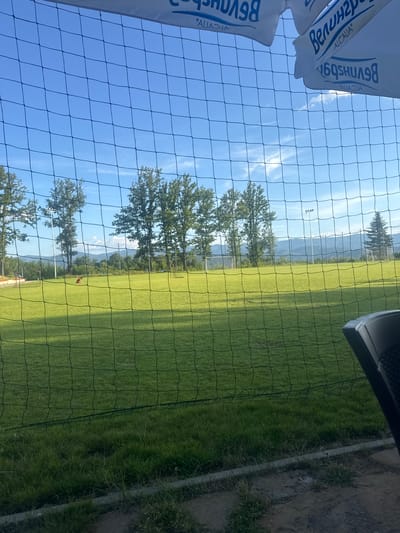 The image depicts an outdoor scene, likely a sports field, viewed through a black mesh net. In the background, a lush green field is visible with several tall trees lining the perimeter. Beyond the field, rolling hills and mountains form a scenic backdrop under a clear blue sky with scattered white clouds. Two soccer goals are present on the field, and a person in an orange shirt is visible kneeling on the grass near one of the goals. The foreground shows the mesh net in sharp focus, with some ground and vegetation underneath. In the upper part of the image, parts of a white banner with blue text are visible, suggesting this is a venue with some branding. The overall impression is of a sunny day in a picturesque, possibly rural, location, with athletic activities taking place. The specific location in Gabrovo, Bulgaria is not evident from the visual information alone.